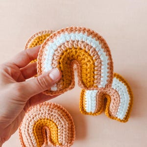 May include: Three crocheted rainbows in varying sizes. The rainbows feature stripes of peach, mustard yellow, and white yarn. The craftsmanship is detailed and the colors are soft and muted, creating a charming and playful aesthetic. These handmade items are perfect for nursery decor or as a unique gift.