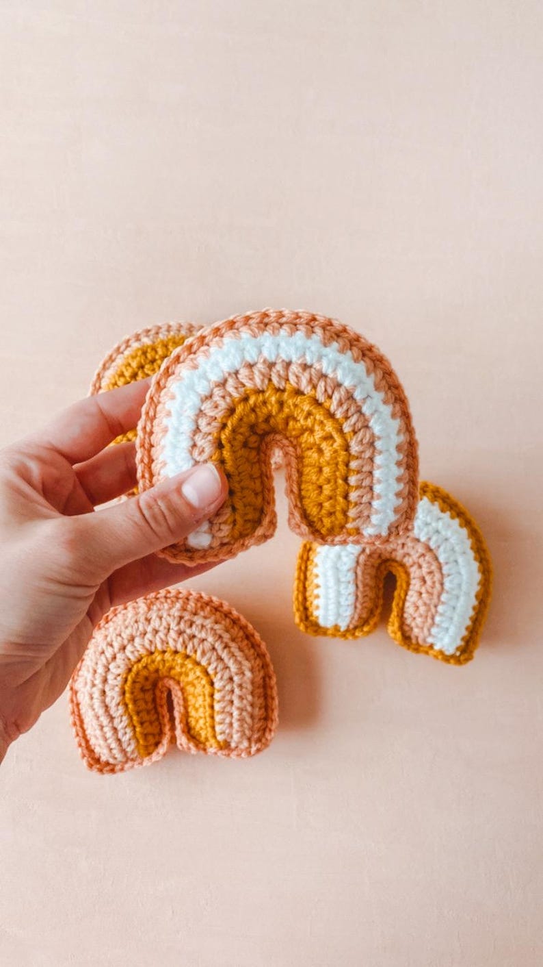 May include: Three crocheted rainbows in varying sizes. The rainbows feature stripes of peach, mustard yellow, and white yarn. The craftsmanship is detailed and the colors are soft and muted, creating a charming and playful aesthetic. These handmade items are perfect for nursery decor or as a unique gift.