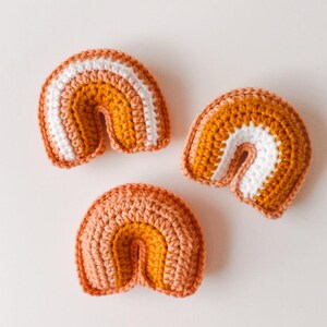 May include: Three small crocheted rainbows in varying shades of orange and mustard yellow with white accents.  Each rainbow is crafted from a chunky yarn, creating a textured appearance. The rainbows are arranged on a plain white background.