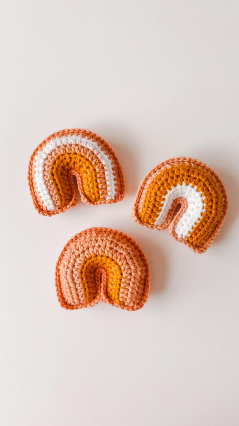 May include: Three small crocheted rainbows in varying shades of orange and mustard yellow with white accents.  Each rainbow is crafted from a chunky yarn, creating a textured appearance. The rainbows are arranged on a plain white background.