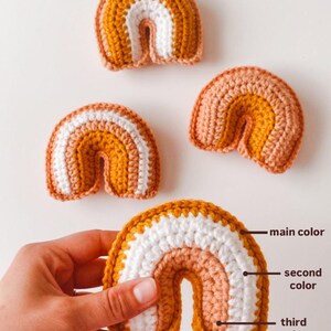 May include: Three crocheted rainbows in varying sizes. The largest rainbow has a mustard yellow top layer, a white middle layer, and a light peach bottom layer.  The other two rainbows use the same color pattern. The image shows the color progression of the yarn used to create the rainbows: main color (mustard yellow), second color (white), and third color (light peach).