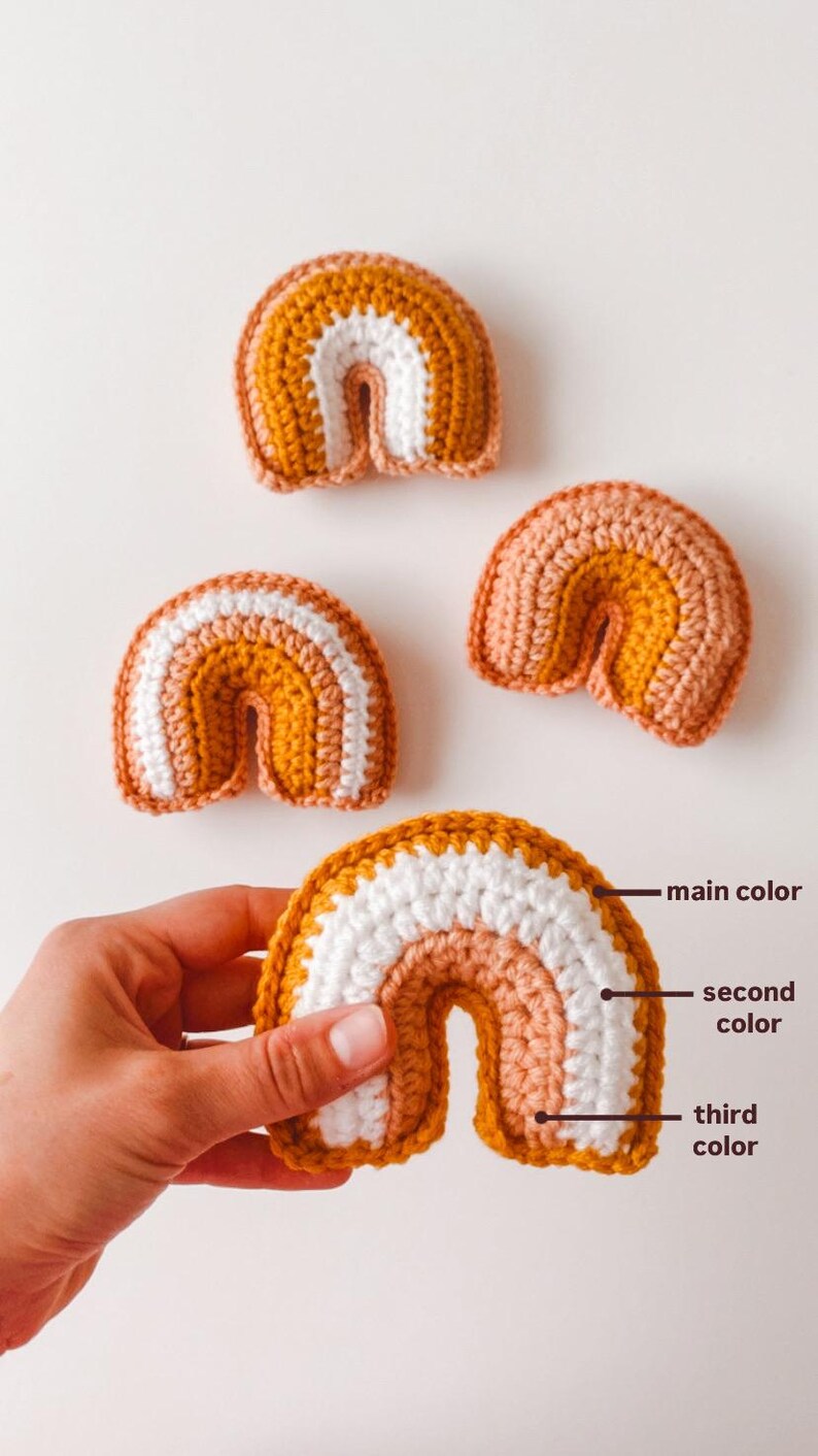 May include: Three crocheted rainbows in varying sizes. The largest rainbow has a mustard yellow top layer, a white middle layer, and a light peach bottom layer.  The other two rainbows use the same color pattern. The image shows the color progression of the yarn used to create the rainbows: main color (mustard yellow), second color (white), and third color (light peach).