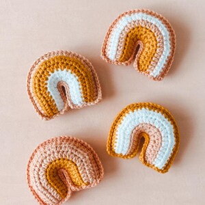May include: Four small crocheted rainbows in varying shades of peach, mustard yellow, and white.  Each rainbow is a handmade, three-toned, amigurumi-style toy. The rainbows are arranged on a light pink background.