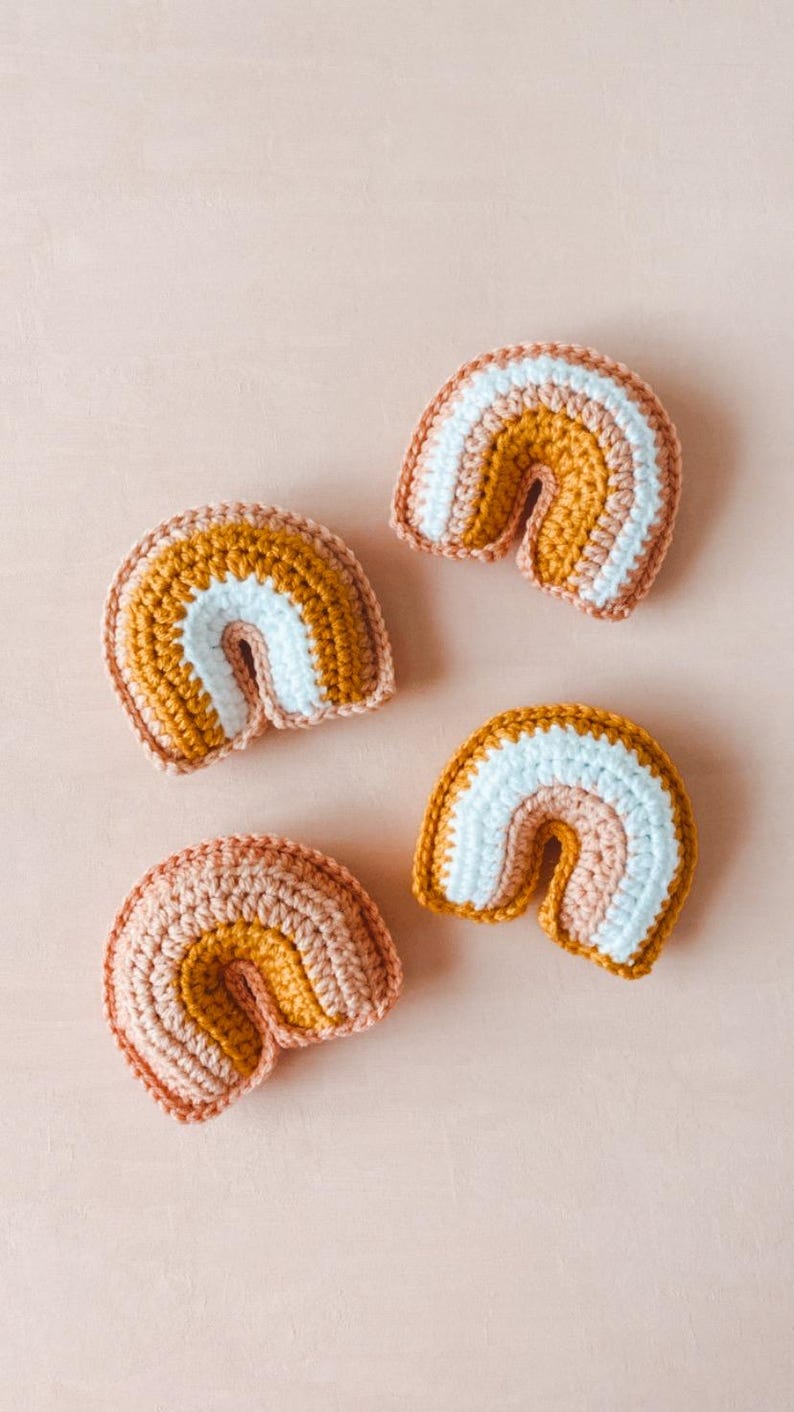 May include: Four small crocheted rainbows in varying shades of peach, mustard yellow, and white.  Each rainbow is a handmade, three-toned, amigurumi-style toy. The rainbows are arranged on a light pink background.
