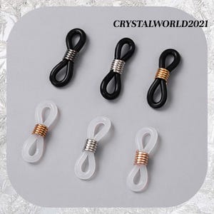 May include: Six eyeglass retainers in black and white, with silver and gold-coloured metal coils. The retainers are arranged on a light grey surface. The text "CRYSTALWORLD2021" is visible in the upper right corner.
