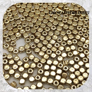 May include: A large quantity of gold-coloured square beads with a hole in the centre. The beads are scattered on a white surface.