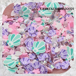 Pode incluir: Uma mistura de enfeites coloridos de argila polimérica, incluindo formas de conchas, flores e corações. A paleta de cores inclui rosa, roxo, branco e verde-azulado, com detalhes em cristal transparente. O texto "CRYSTALWORLD2021" é visível.