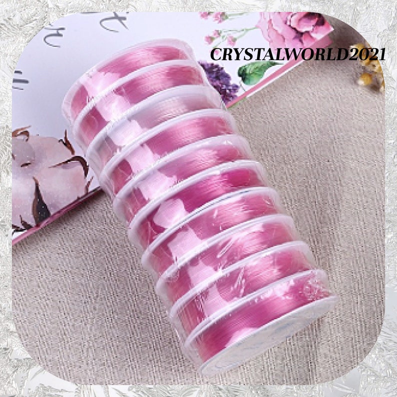 May include: A close-up of a stack of pink elastic cord spools. The spools are wrapped in clear plastic and have a white label with the text "CRYSTALWORLD2021".