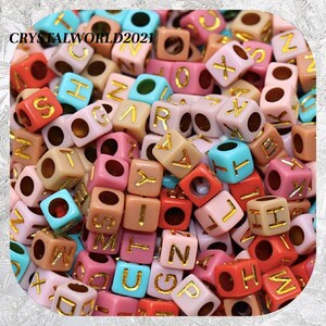 May include: A large pile of colorful plastic letter beads. The beads are in various colors, including pink, blue, red, and brown. Each bead has a gold letter on it. The letters are all different and are in a random order.