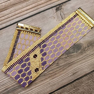 May include: A beaded bracelet with a gold clasp and a geometric pattern. The bracelet features a combination of gold, purple, and lavender beads. The bracelet is approximately 1 inch wide.