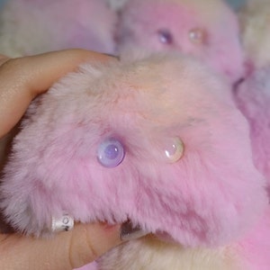 Op de afbeelding: Een pluche roze en witte knuffel met iriserende ogen. Het speelgoed is gemaakt van zacht, pluche stof en heeft een speelse, wolkachtige uitstraling.