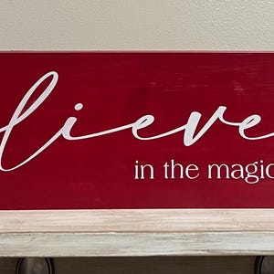 Op de afbeelding: Een rechthoekig, rood houten bord met het woord "believe" in witte cursieve letters. Daaronder staat de tekst "in the magic of CHRISTMAS" in witte schreefloze letters. Het bord staat op een witte plank.