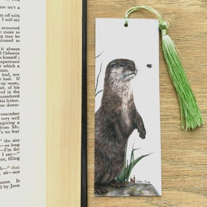 May include: A watercolour bookmark featuring a detailed illustration of an otter standing on a rock with green grass and a bee flying nearby. The bookmark has a green tassel.