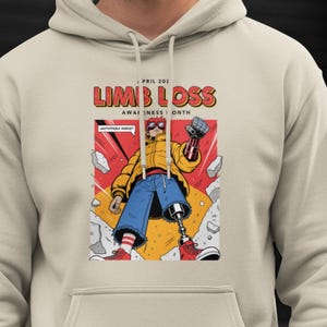 Puede incluir: Sudadera con capucha beige con un gráfico frontal que anuncia el "Mes de Concienciación sobre la Pérdida de Extremidades". El diseño, estilo cómic, muestra a una persona con una prótesis de pierna, chaqueta amarilla, vaqueros azules y zapatillas rojas.