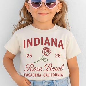 May include: A child wearing a cream-colored t-shirt with the text "INDIANA" in maroon, along with "Rose Bowl" and "Pasadena, California." A rose graphic is between the text. The child is also wearing pink heart-shaped sunglasses and blue jeans.