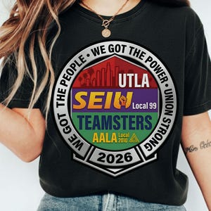 Op de afbeelding: Zwart T-shirt met een cirkelvormige afbeelding. Het ontwerp bevat de tekst "WE GOT THE PEOPLE" en "WE GOT THE POWER", evenals "UTLA", "SEIU", "TEAMSTERS", "AALA 2010" en het jaar "2026".