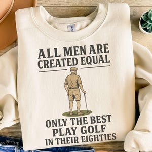 Puede incluir: Sudadera color crema con el texto "ALL MEN ARE CREATED EQUAL" y "ONLY THE BEST PLAY GOLF IN THEIR EIGHTIES." Un gráfico de un golfista está debajo del texto. La sudadera se combina con jeans azules y un sombrero marrón.