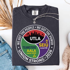 Op de afbeelding: Donkergrijze sweatshirt met een cirkelvormige afbeelding. De afbeelding bevat de tekst "WE GOT THE PEOPLE WE GOT THE POWER" en "UNION STRONG 2026", samen met logo's van UTLA, Teamsters AALA en SEIU Local 99. Een zilveren ketting en jeans zijn ook zichtbaar.
