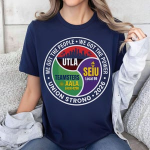 Op de afbeelding: Marineblauw T-shirt met een cirkelvormig ontwerp. Het ontwerp bevat de tekst "WE GOT THE PEOPLE WE GOT THE POWER" en "UNION STRONG 2026". Logo's van UTLA, Teamsters AALA en SEIU Local 99 zijn ook aanwezig.