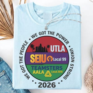 Op de afbeelding: Lichtblauw T-shirt met een cirkelvormig ontwerp. Het ontwerp bevat een silhouet van een stadsgezicht, de tekst "WE GOT THE POWER" en de woorden "UTLA", "SEIU Local 99" en "TEAMSTERS AALA Local 2010". Het jaar "2026" is ook aanwezig.