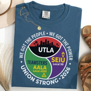 Op de afbeelding: Een blauw t-shirt met een cirkelvormige afbeelding met de tekst "WE GOT THE PEOPLE WE GOT THE POWER UNION STRONG 2026." De afbeelding bevat de logo's van UTLA, Teamsters AALA en SEIU Local 99. Een gouden ketting en sandalen zijn ook zichtbaar.