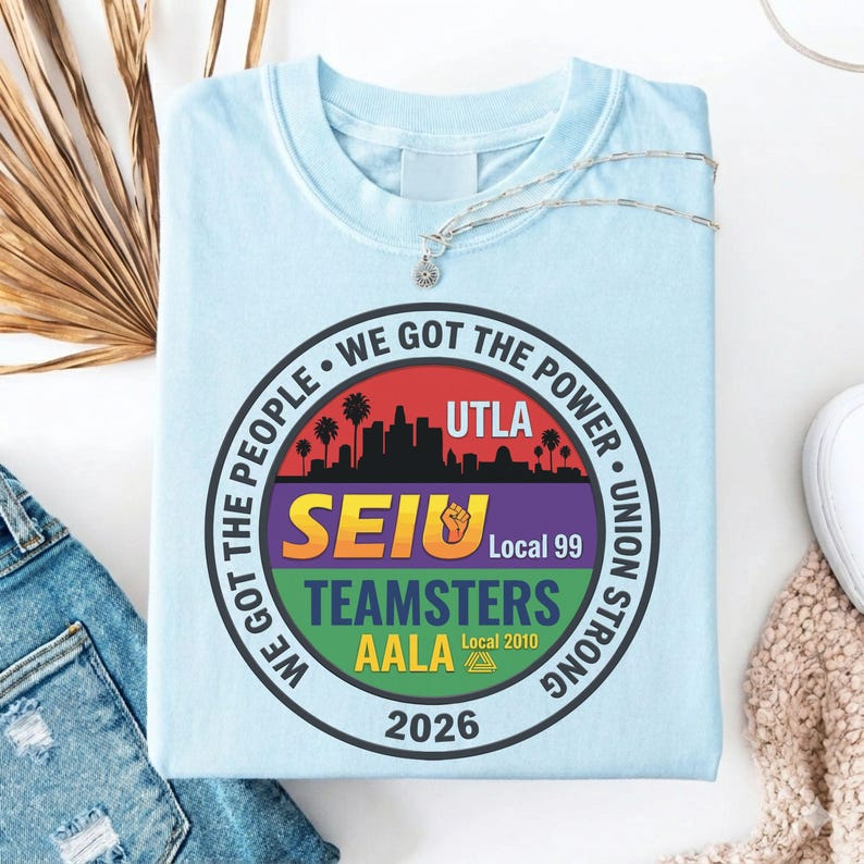 K&ouml;nnte beinhalten: Hellblaues T-Shirt mit einem kreisf&ouml;rmigen Grafikdesign. Das Design zeigt eine Stadtsilhouette, Palmen und den Text "WE GOT THE POWER" und "UNION STRONG". Es enth&auml;lt auch "SEIU", "TEAMSTERS" und "AALA" mit dem Jahr "2026".