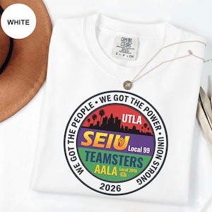 K&ouml;nnte beinhalten: Wei&szlig;es T-Shirt mit einem kreisf&ouml;rmigen Grafikdesign mit dem Text "WE GOT THE PEOPLE" und "WE GOT THE POWER". Die Grafik enth&auml;lt die Texte "UTLA", "SEIU", "TEAMSTERS" und "AALA". Das Shirt hat auch den Text "2026".