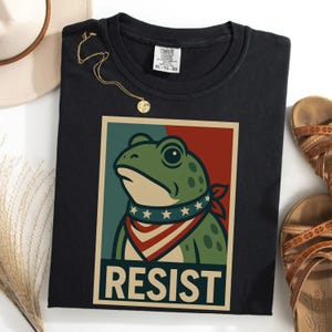Op de afbeelding: Zwart T-shirt met een kikker-afbeelding met een rood, wit en blauw bandana. De kikker staat tegen een rode en blauwe achtergrond met het woord "RESIST". Een gouden ketting en sandalen zijn ook zichtbaar.