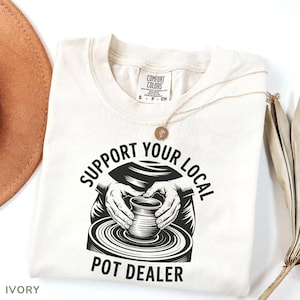 May include: Ivory-colored t-shirt with the text "SUPPORT YOUR LOCAL POT DEALER" in a black, stylized font. The graphic depicts hands shaping a pot on a pottery wheel. A gold necklace with a pendant rests on the shirt.