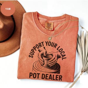 May include: A coral-coloured t-shirt with the text "SUPPORT YOUR LOCAL POT DEALER" in black. The graphic depicts hands shaping a pot on a pottery wheel. A gold necklace and a brown hat are also visible.