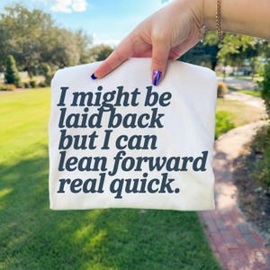 May include: A white t-shirt with the text "I might be laid back but I can lean forward real quick" in dark blue lettering. The shirt is held up against a background of green grass and trees.