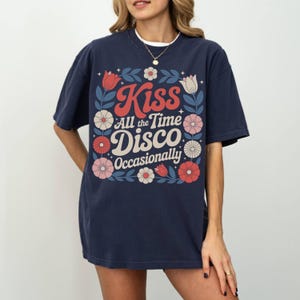 May include: Navy blue t-shirt with the text "Kiss All the Time Disco Occasionally" in a retro font, surrounded by floral and leaf illustrations in red, white, and blue. The shirt has short sleeves and a relaxed fit.