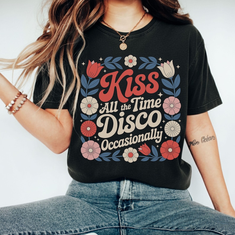 May include: Black t-shirt with a floral design and the words "Kiss All the Time Disco Occasionally" in a retro font. The design includes red, pink, and white flowers with blue leaves. The shirt is a casual wear item.