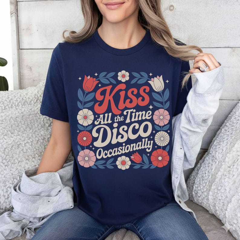 May include: Navy blue t-shirt with a floral design and the words "Kiss All the Time Disco Occasionally" in a retro font. The design includes red, white, and pink flowers and green leaves. The shirt is a short-sleeved crew neck.