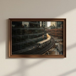 Op de afbeelding: Een ingelijste foto van een stadsgezicht met een trein op rails die door een stadslandschap slingert. De afbeelding staat in een bruine houten lijst en hangt aan een witte muur. De gebouwen zijn een mix van modern glas en oudere bakstenen structuren.