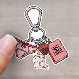 May include: A keychain with a silver clasp holding several charms. Charms include a pair of pink underwear, a chocolate-covered strawberry, a candle, and a book with the text "SMUT LOVER" on the cover.