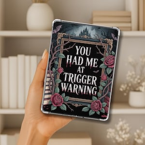 May include: A black tablet case with the text "YOU HAD ME AT TRIGGER WARNING" in white. The case features a gothic design with roses, bats, and a castle illustration. The case is held in a person's hand.