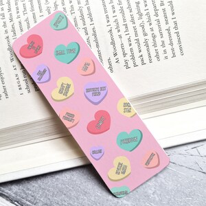 May include: Pink bookmark featuring a pattern of colourful candy hearts. Each heart has a different message, such as "Best Friend", "College", "Rock Star", and "Sweetest Smile".