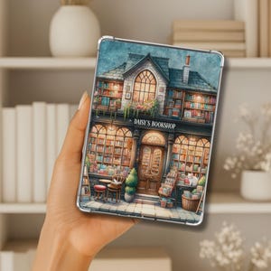 May include: A clear tablet case with a watercolour illustration of "Daisy's Bookshop." The image shows a charming bookstore with a detailed facade, filled with books, and warm lighting. The case is held in a person's hand, showcasing the design.