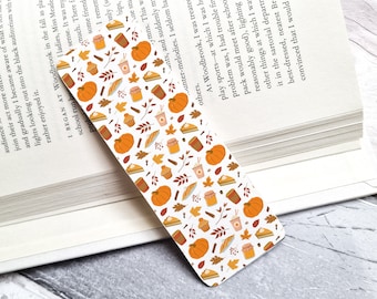 May include: A white bookmark with a fall-themed pattern. The bookmark features orange pumpkins, brown leaves, cinnamon sticks, coffee cups, and pie slices.