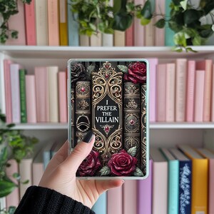 May include: A clear phone case with a black and gold design featuring a stack of antique books with red roses and the text "I Prefer the Villain".