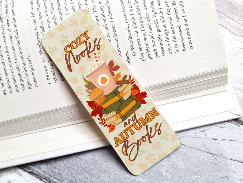 Puede incluir: Un marcap&aacute;ginas con tem&aacute;tica oto&ntilde;al que presenta una pila de libros, una taza de caf&eacute; y hojas de oto&ntilde;o. El marcap&aacute;ginas es beige con texto marr&oacute;n que dice "Cozy Nooks and Autumn Books".