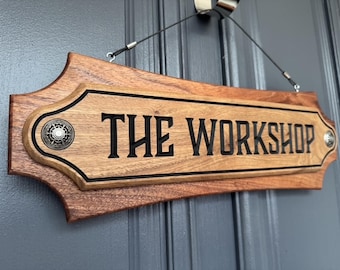 May include: A rectangular wooden sign with beveled edges and a natural wood grain finish. The text THE WORKSHOP is carved in black, bold, sans-serif capital letters. The sign features a decorative silver-toned metal stud on each side and hangs.