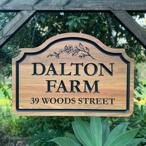 May include: A rectangular wooden sign with a curved top edge, featuring the text DALTON FARM and 39 WOODS STREET in black carved lettering. A black floral illustration is centred above the text. The sign is suspended within a dark wood frame.