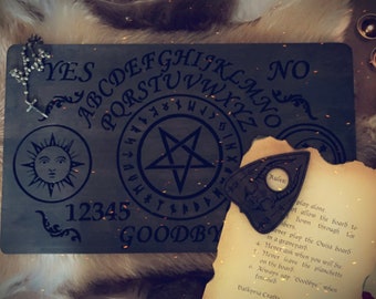 May include: A dark wooden spirit board featuring the alphabet, numbers 1-5, a sun icon, a pentagram, and the word GOODBYE. A matching dark wooden planchette rests on a piece of parchment paper printed with rules, alongside a small silver rosary.