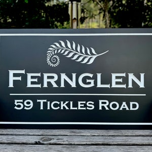 May include: A rectangular black sign with white lettering and a white border. The sign reads "FERNGLEN" above "59 TICKLES ROAD". A white fern design is at the top. The sign is displayed outdoors.
