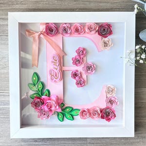 Puede incluir: Una obra de arte en caja de sombra enmarcada en blanco con la letra "E" rosa adornada con flores de papel en tonos rosas y rojos. La letra está adornada con una cinta rosa y el nombre "Evie" en purpurina dorada. Hojas verdes añaden contraste.