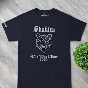 Puede incluir: Una camiseta negra de manga corta con un gráfico plateado de una cabeza de lobo geométrica. El texto Shakira aparece sobre el lobo y #LMYNLWorldTour 2026 debajo. La etiqueta interior dice Made With L y la palabra design está en la manga.