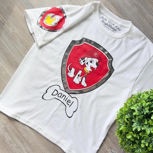Puede incluir: Camiseta blanca con un bombero dálmata de dibujos animados sobre un escudo rojo y plateado. La camiseta incluye el nombre "Daniel" en un gráfico con forma de hueso. Un emblema de fuego está en la manga. El texto "Made With Love SinghiDesign" está impreso en el cuello.