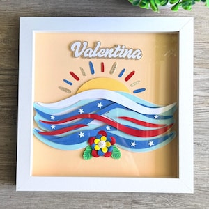 May include: A framed paper art piece featuring the name "Valentina" in gold glitter lettering. The artwork depicts a yellow sun, blue and red waves with white stars, and a colorful flower. The frame is white.